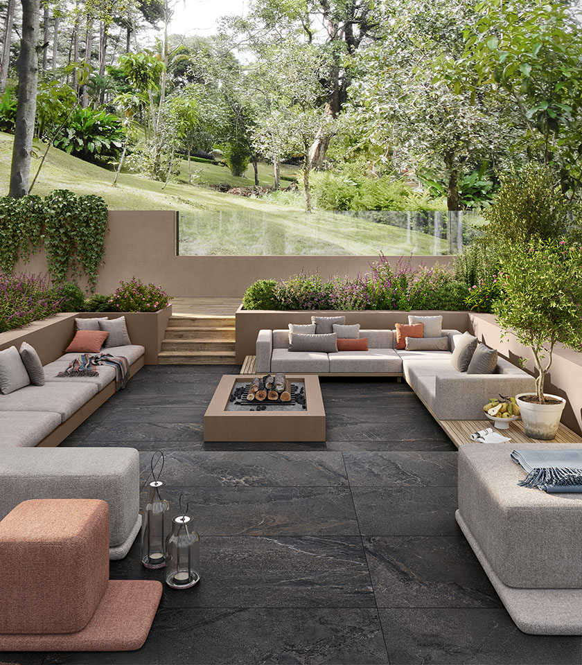 Zona living outdoor immersa nel verde con pavimento della collezione Anthology effetto pietra nella tonalità Dark Rough ed elementi della collezione Pigmenti nella tonalità Pumice.