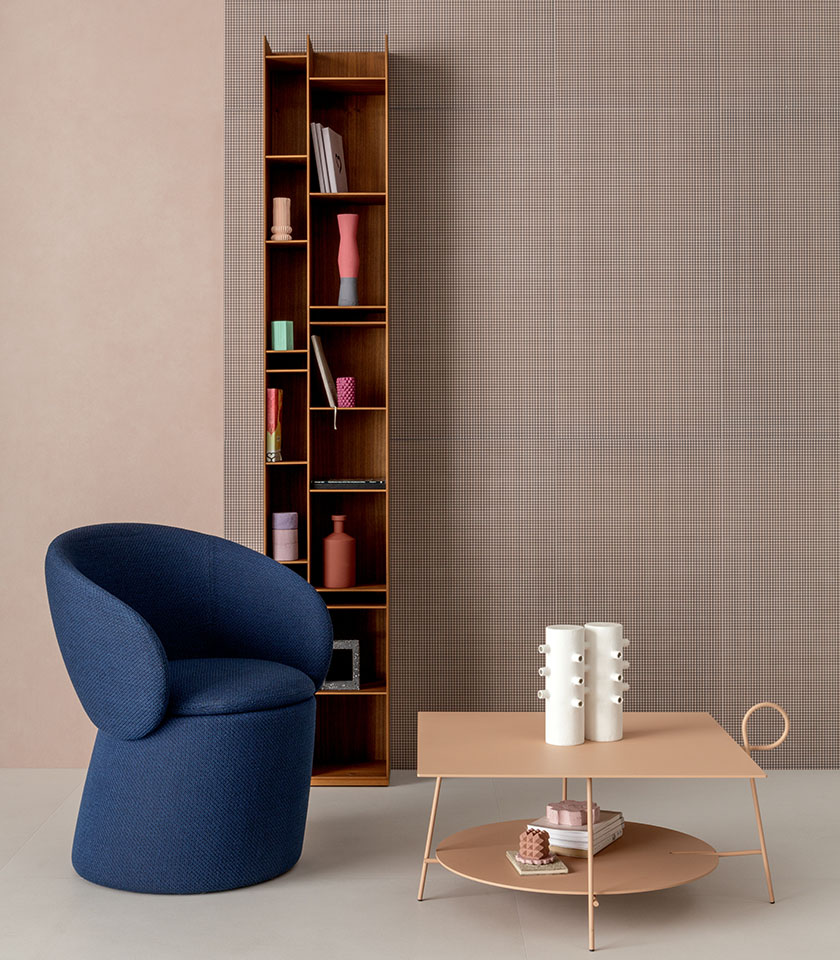 Interno contemporaneo con poltrona blu, tavolino dalle tonalità cipria, libreria in legno e parete decorativa a microfantasia.