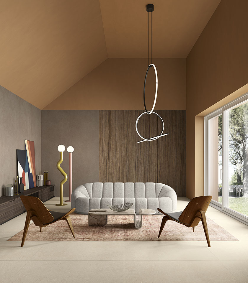 Soggiorno contemporaneo con pavimento beige effetto pietra, divano scultoreo, poltrone in legno e grandi vetrate affacciate sul verde.