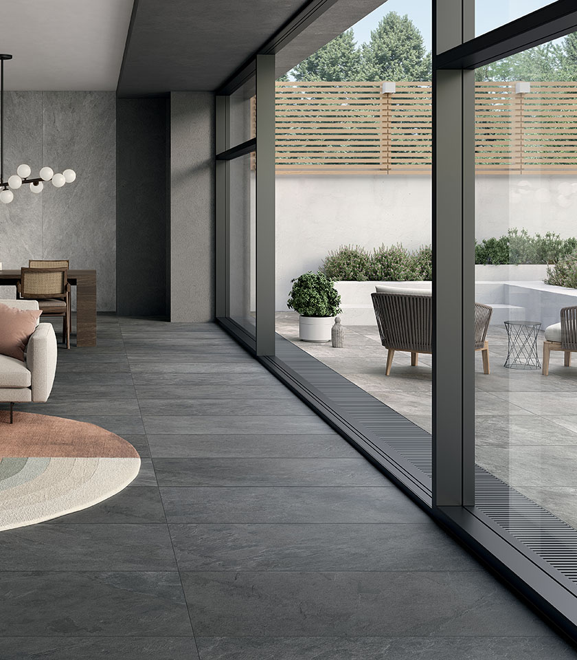 Ambiente contemporaneo con pavimentazione effetto pietra grigia in continuità tra interno ed esterno, ampie vetrate e terrazza arredata dal design essenziale.