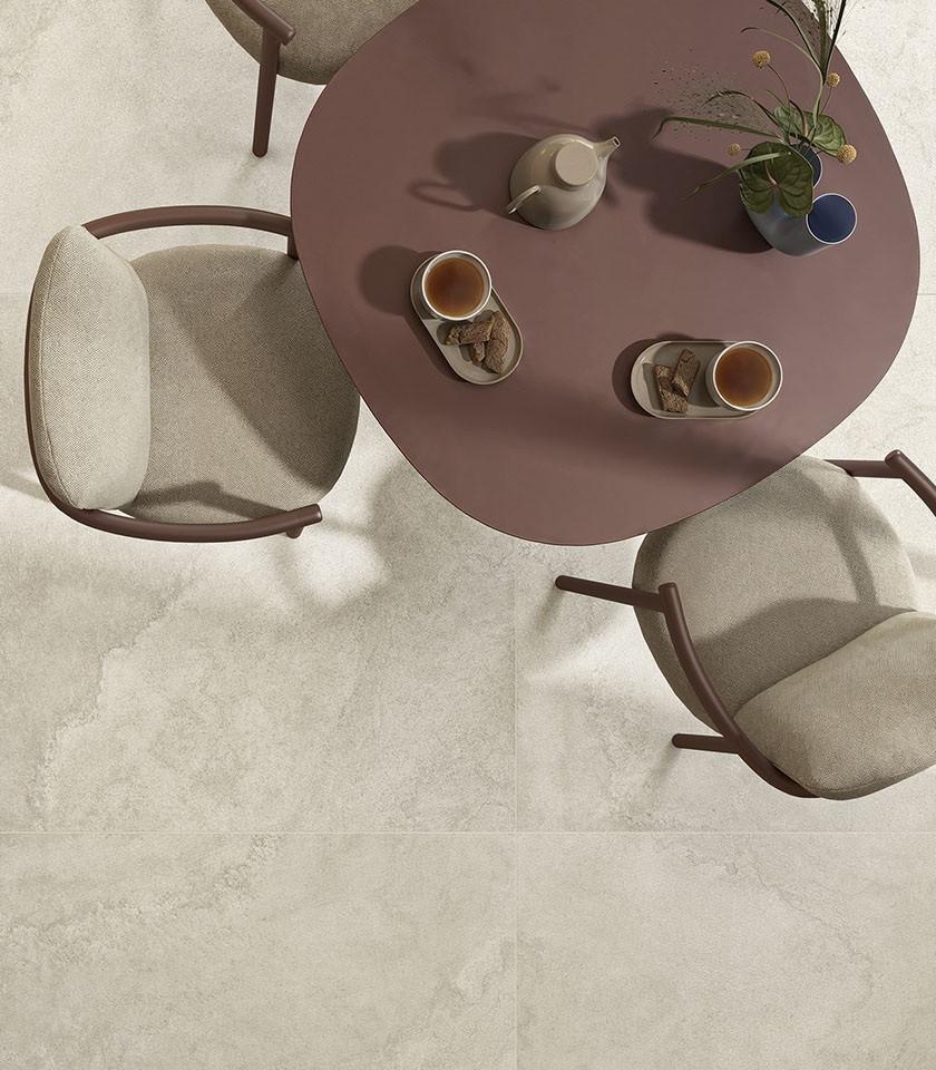 Tavolo rotondo color terracotta con servizio da tè e sedie imbottite beige su pavimento della collezione Pulse, effetto pietra nella tonalità Shell.