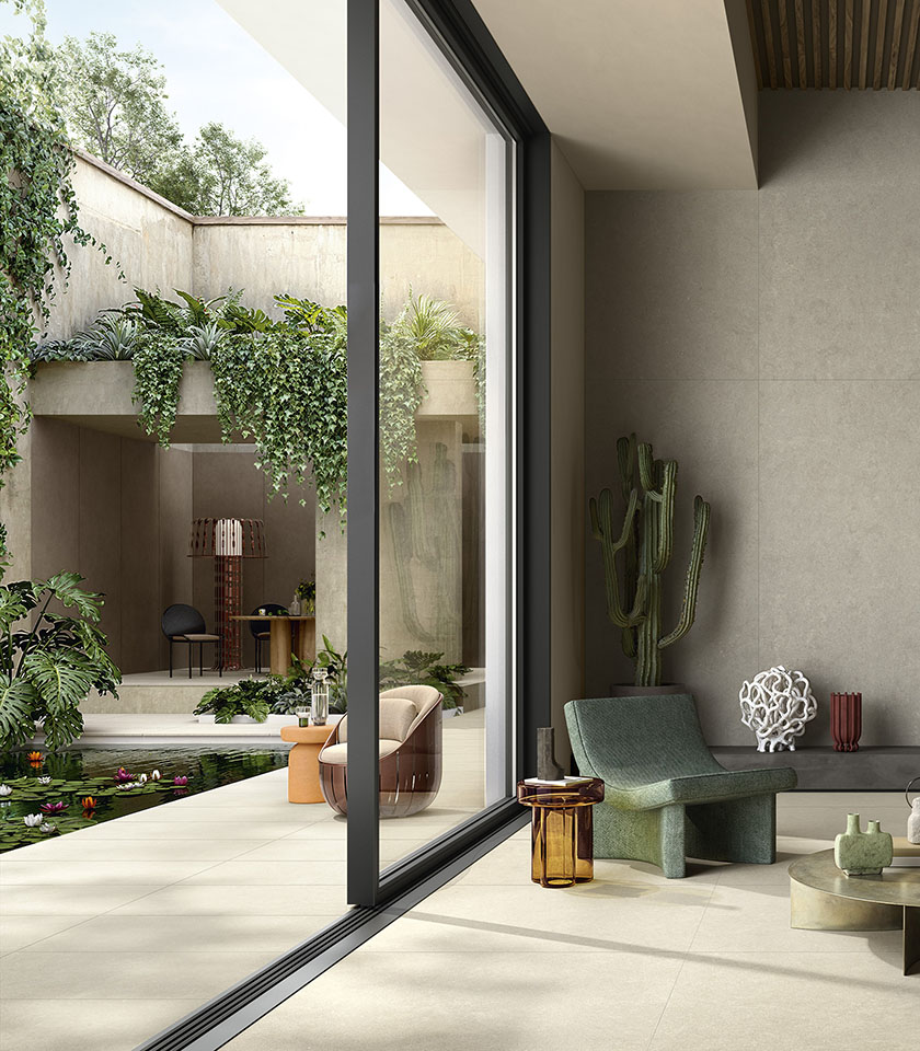 Ambiente indoor-outdoor con pavimento in gres porcellanato chiaro, ampie vetrate, vegetazione rigogliosa e arredi contemporanei.