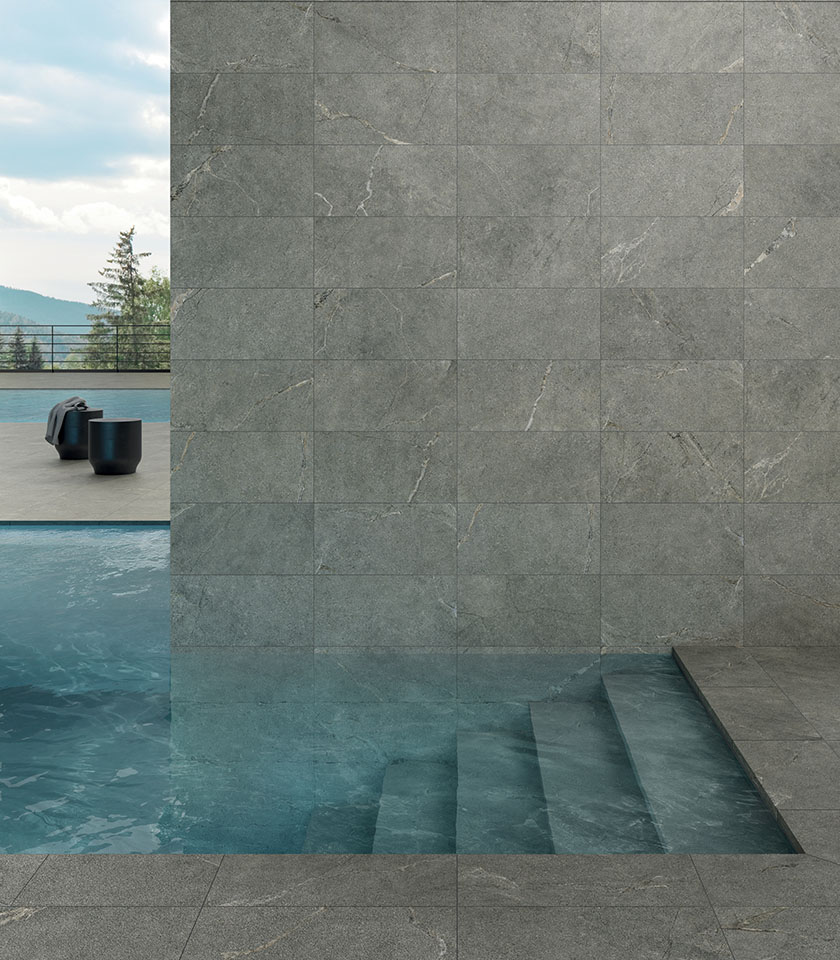 Piscina con rivestimento e pavimentazione in gres porcellanato effetto pietra grigia, gradini immersi e vista sulle montagne.