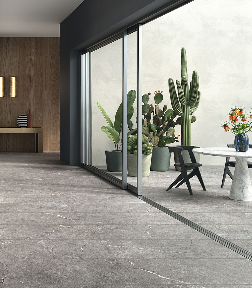 Ambiente contemporaneo con pavimento effetto pietra grigia in continuità tra interno ed esterno, ampie vetrate, arredi essenziali e piante decorative.