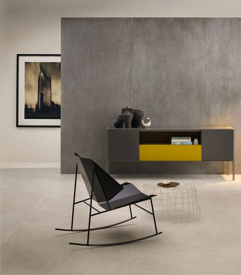 Ambiente living minimal chic con pavimento chiaro effetto cemento, rivestimento grigio a grande formato, arredi essenziali e dettagli di design contemporaneo.
