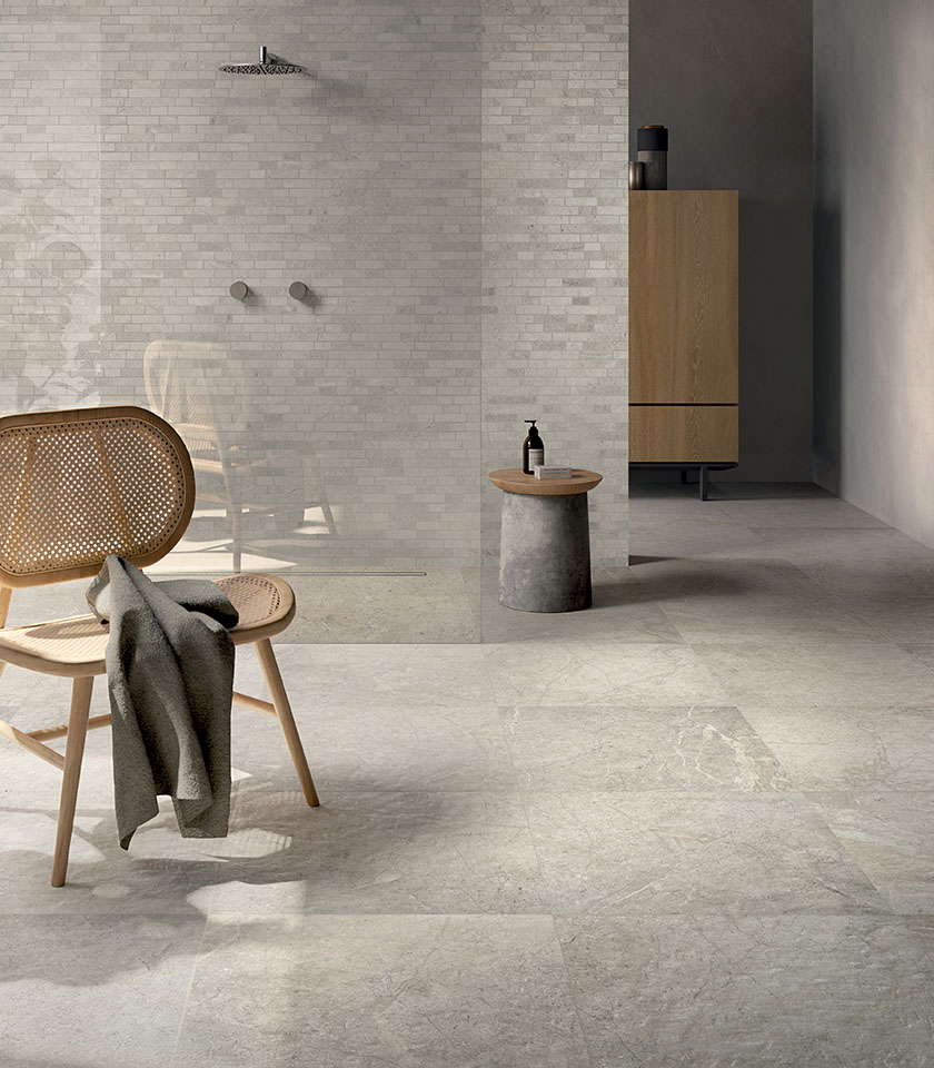 Bagno minimal chic con pavimento effetto pietra chiara, rivestimento materico tono su tono, doccia walk-in e arredi in legno dal design essenziale.