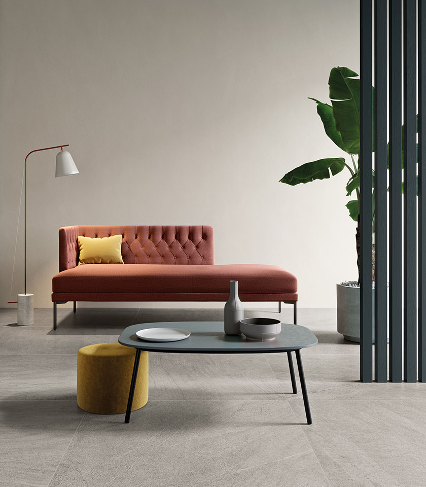 Living minimal chic con pavimento effetto pietra grigia, chaise longue color terracotta, arredi essenziali e accenti caldi dal design contemporaneo.